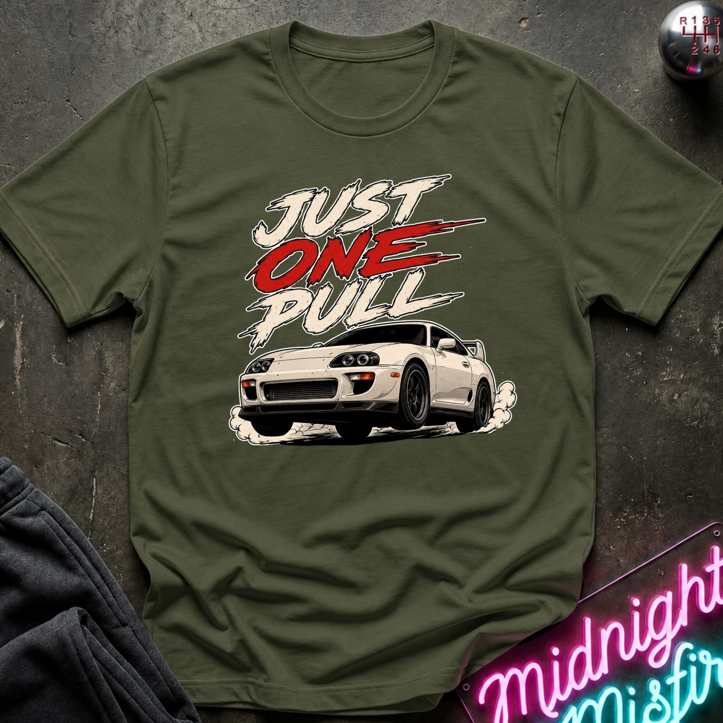 An olive green graphic t-shirt featuring a white sports car and the text 'JUST ONE PULL' in white and red, styled flat on a dark textured background with a neon sign and dark pants partially visible.