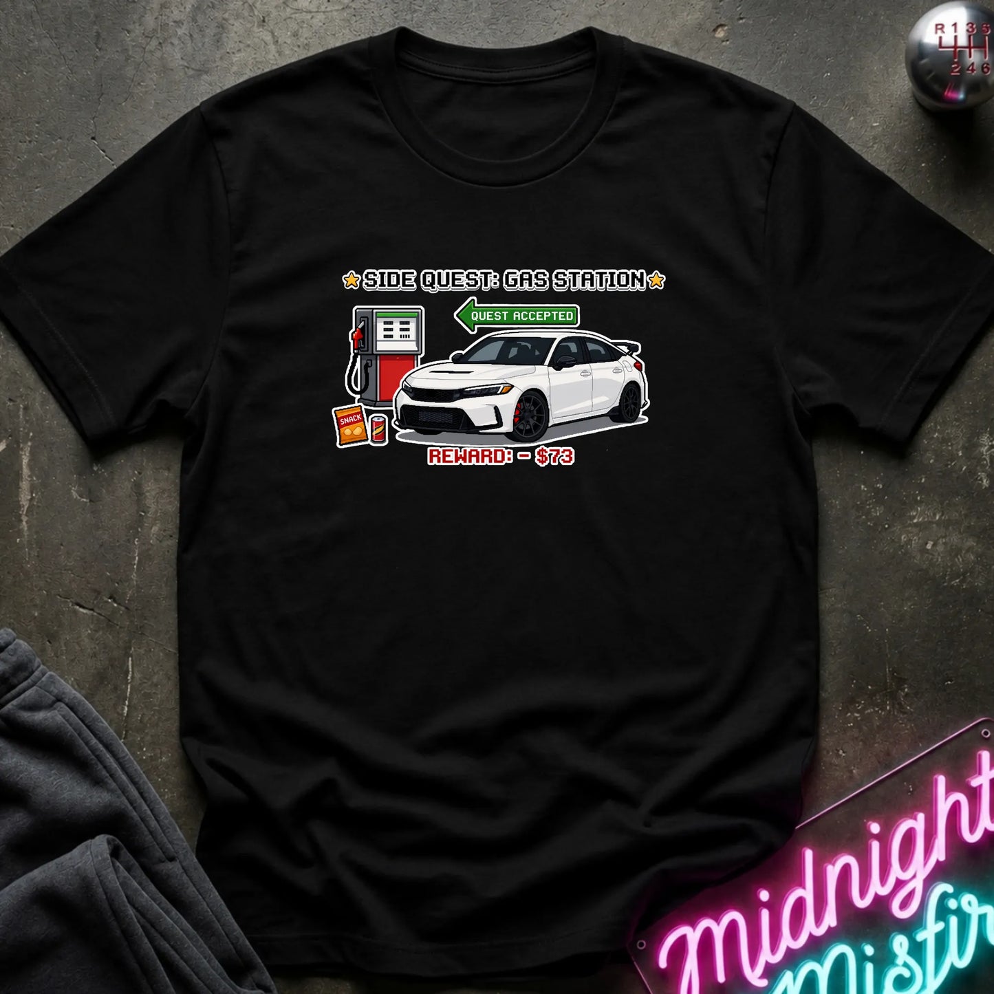 A black short-sleeve graphic t-shirt laid flat on a dark textured surface. The graphic features a white sports car, a red and white gas pump, and text reading 'SIDE QUEST GAS STATION'.