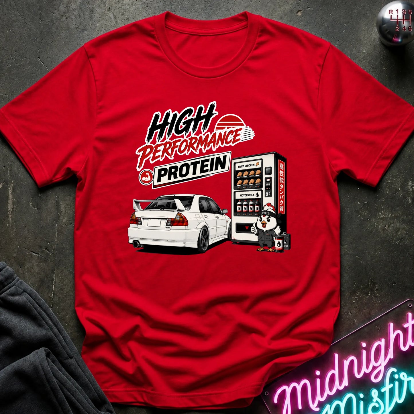 A red short-sleeve graphic T-shirt laid flat on a dark textured surface. The shirt features a white car and a vending machine illustration with the text 'HIGH PERFORMANCE PROTEIN' in stylized lettering.