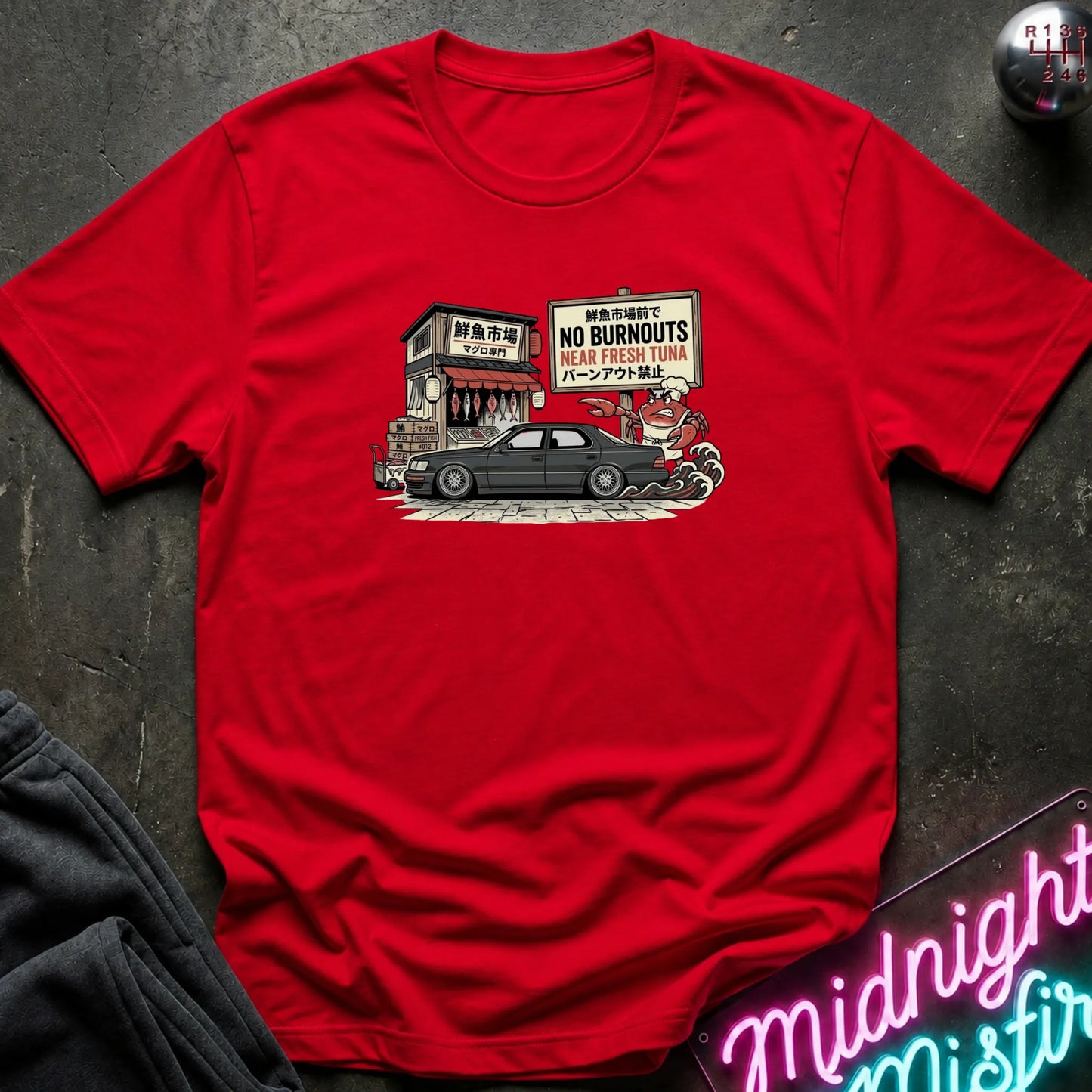 A red short-sleeve crewneck t-shirt laid flat on a dark textured surface. The shirt features a central graphic illustration of a black car with a sign that reads 'NO BURNOUTS HEAD FRESH TUNA'.