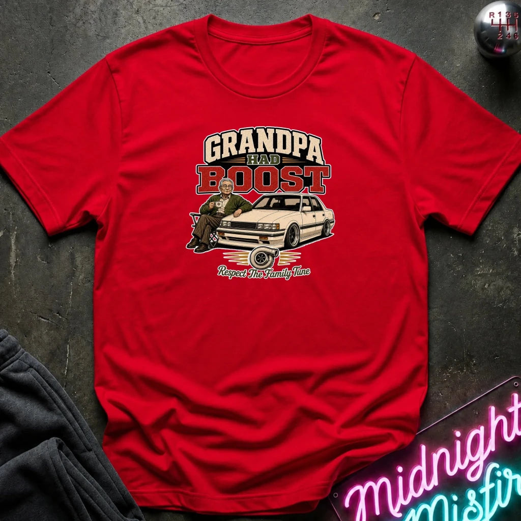 red short-sleeve graphic t-shirt laid flat on a dark textured surface. The graphic features the text 'GRANDPA RAW BOOST' in beige and red, above an illustration of a man sitting in a car with the phrase 'Respect The Family Tune' below.