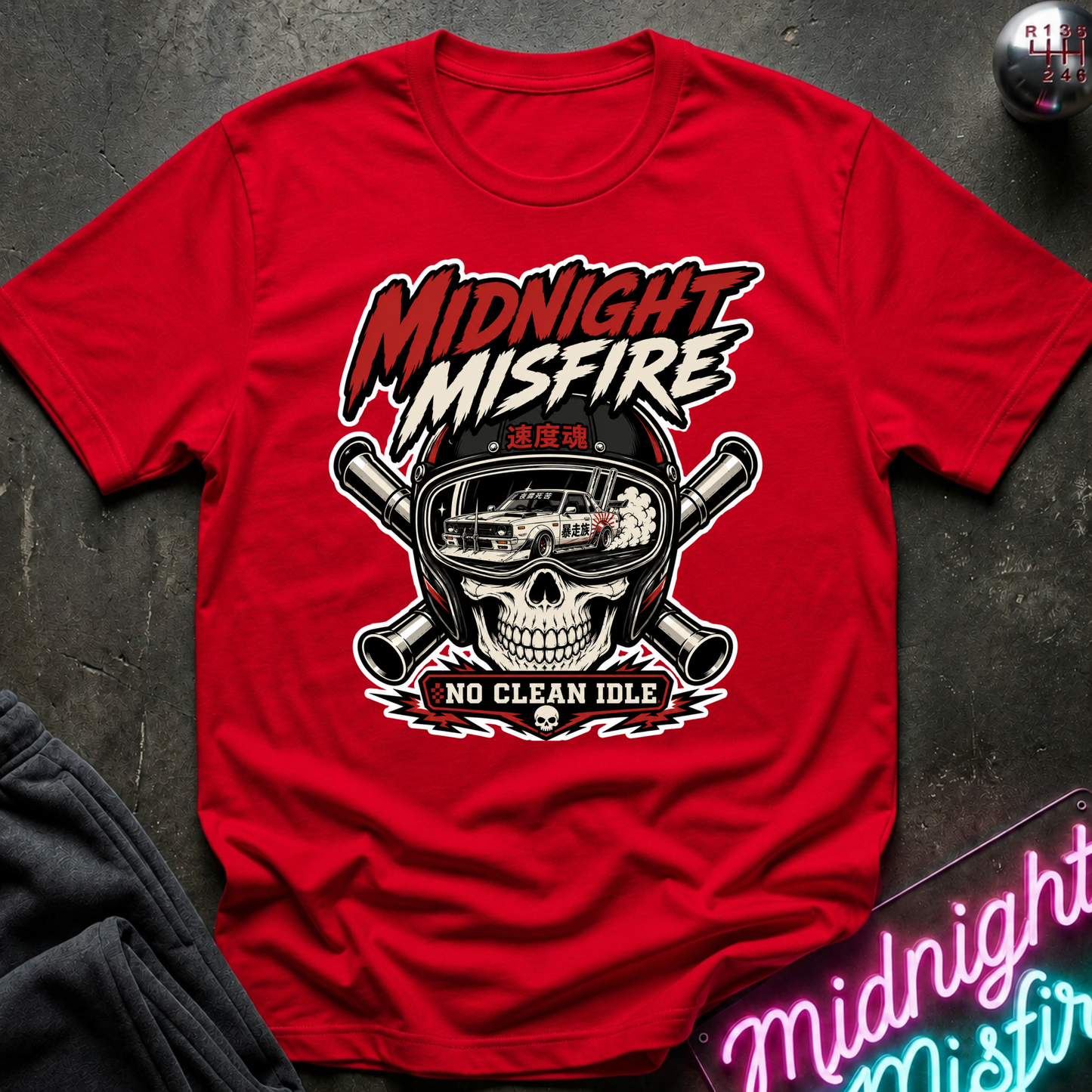 A red short-sleeve graphic T-shirt laid flat on a dark textured surface. The shirt features a large front print with the words “MIDNIGHT MISFIRE” above a skull wearing a helmet with crossed pipes and a car reflected in the visor, and “NO CLEAN IDLE” below.