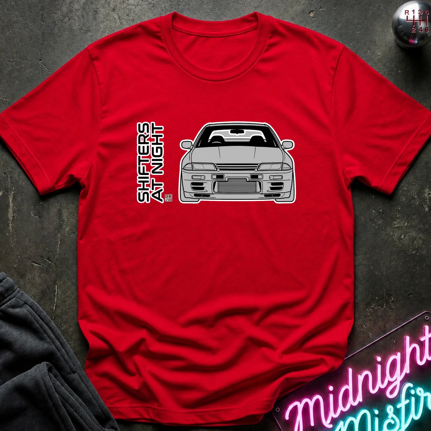 A red short-sleeve graphic T-shirt laid flat on a dark textured surface, featuring a white line drawing of a car and vertical text reading 'SHIFT AT NIGHT' on the chest. A neon sign and dark fabric are partially visible around the shirt as styled props.