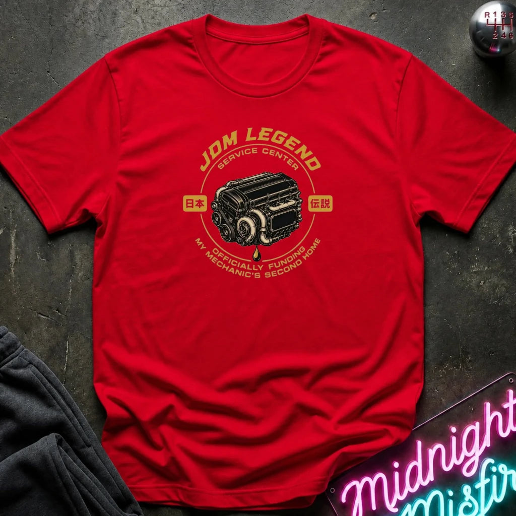 red graphic t-shirt laid flat on a dark textured surface. The shirt features a vintage-style yellow and black engine graphic with the text 'JDM LEGEND' and 'MY SPECIALTY FUNDS MECHANIC'S SECOND HAND'.