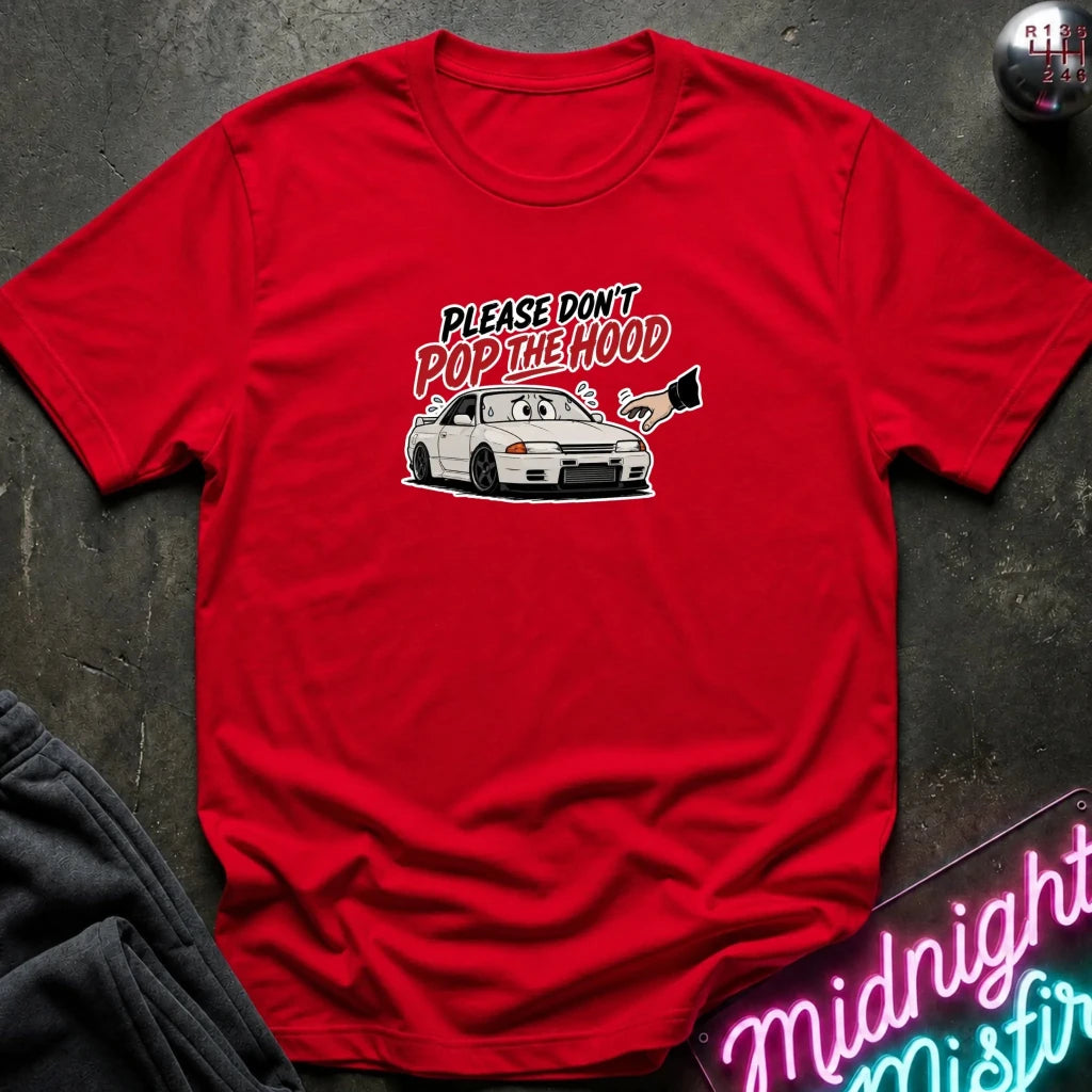A red short-sleeve graphic t-shirt laid flat on a dark textured surface. The graphic features a white sports car with a cartoon face and the text 'PLEASE DON'T POP THE HOOD' in stylized lettering.