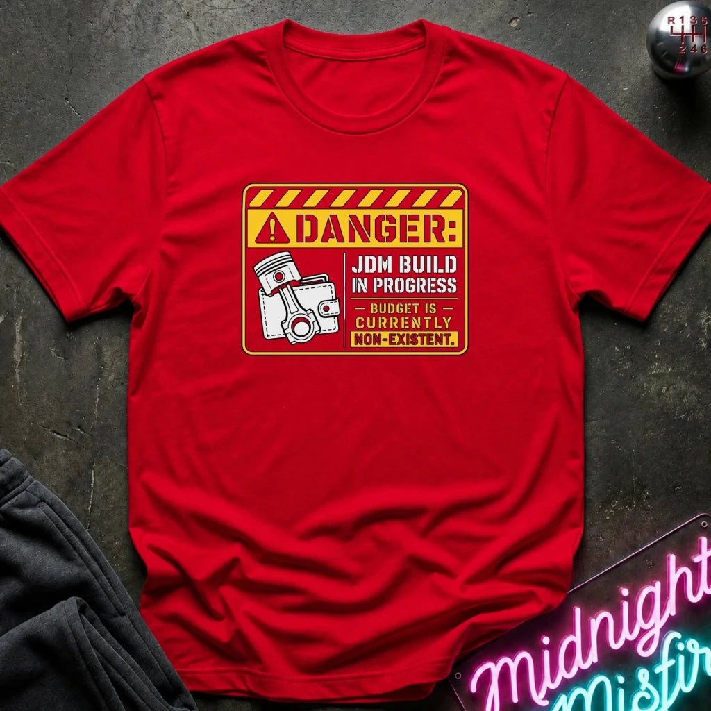 A red short-sleeve crewneck t-shirt laid flat on a dark textured surface. The shirt features a humorous warning label graphic on the chest that reads 'DANGER: JDM BUILD IN PROGRESS BUDGET IS CURRENTLY NON-EXISTENT.
