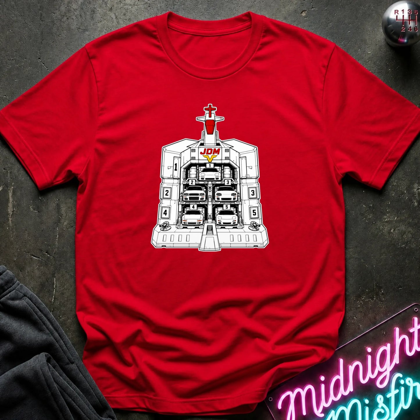 A red short-sleeve crewneck t-shirt laid flat on a dark textured surface, featuring a black and white graphic of the JDM (Japanese Domestic Market) cars in a launch bay.
