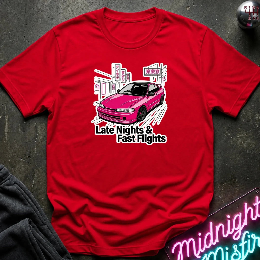 A red short-sleeve graphic T-shirt laid flat on a dark textured surface. The shirt features a pink car illustration with white and black text reading 'Late Nights & Fast Flights.