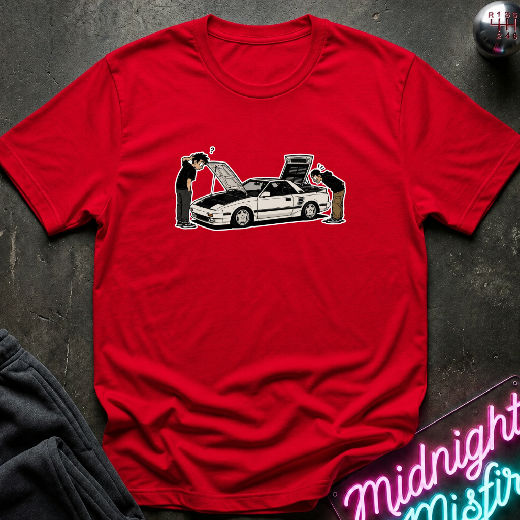 A red short-sleeve crewneck t-shirt laid flat on a dark textured surface, featuring a graphic of a white sports car with its hood and trunk open, flanked by two people inspecting it. A neon sign reading 'Midnight Mistral' is partially visible in the bottom right corner.