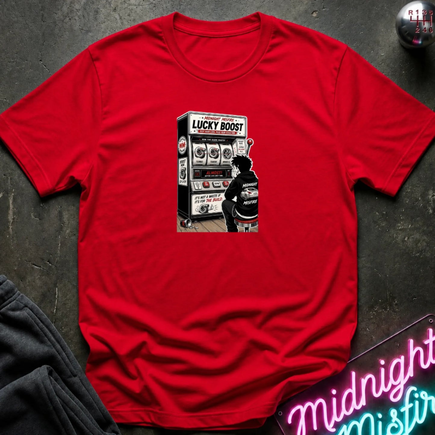 A red  short-sleeve t-shirt with a retro-style graphic of a slot machine, displayed flat on a dark textured surface with a neon sign and sweatpants partially visible.