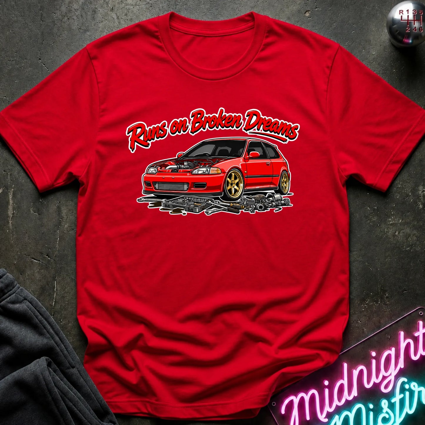 red graphic t-shirt laid flat on a dark textured surface. The shirt features a red sports car with gold wheels and the text 'Runs on Broken Dreams' in red and white.