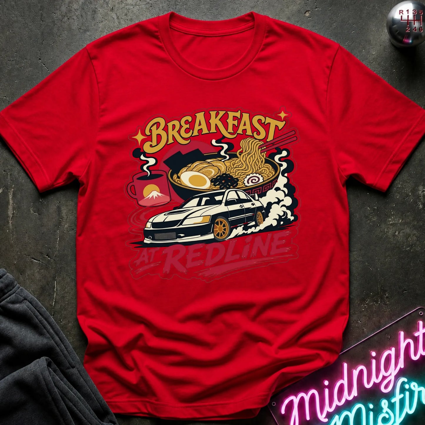 A red short-sleeve graphic T-shirt laid flat on a dark textured surface. The shirt features a colorful retro-style print with the words “Breakfast at Redline,” a white car, ramen noodles, an egg, and red abstract shapes.