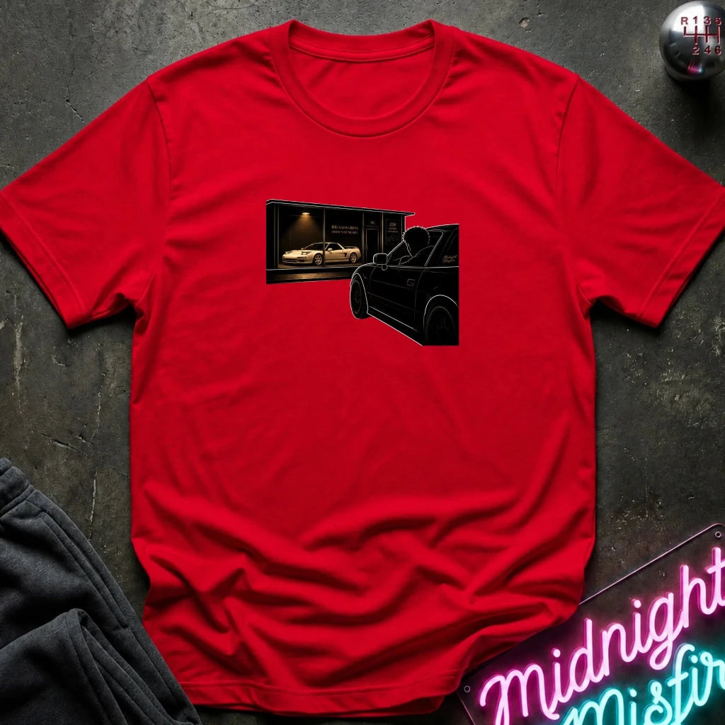 A red short-sleeve crewneck t-shirt laid flat on a dark textured surface, featuring a graphic print of a black car silhouette in front of a garage with a white sports car inside. 