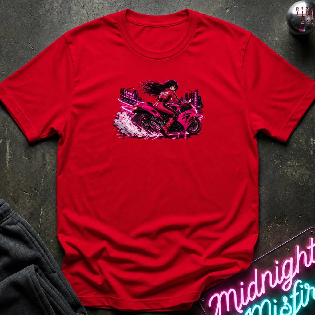 A bright red short-sleeve t-shirt laid flat on a dark textured surface, featuring a central graphic of a woman riding a pink motorcycle with a neon city background. A folded pair of dark pants is visible in the lower-left corner, and a neon sign and a small round object appear at the edges.