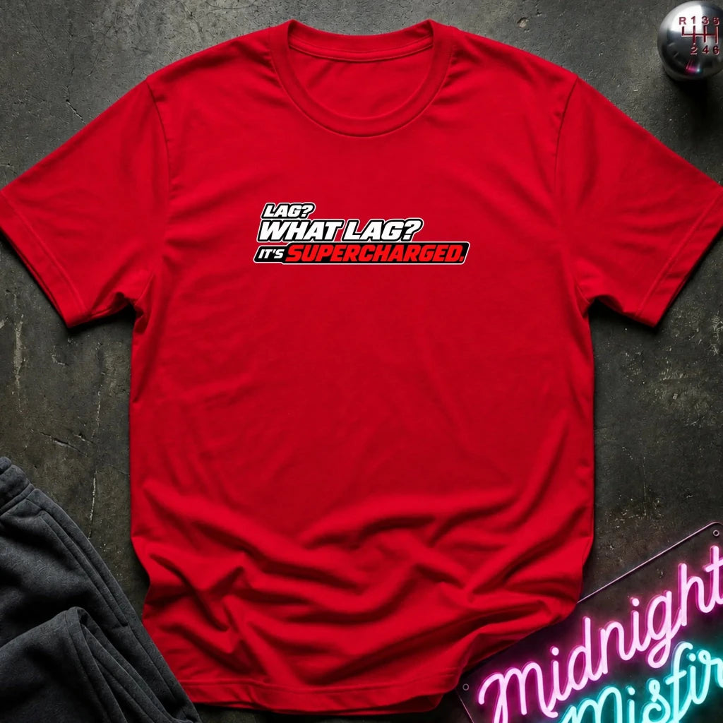 A red short-sleeve crewneck t-shirt with a graphic text print reading 'LAG? WHAT LAG? IT'S SUPERCHARGED' in white and black with a red outline, laid flat on a dark textured surface with lifestyle props.