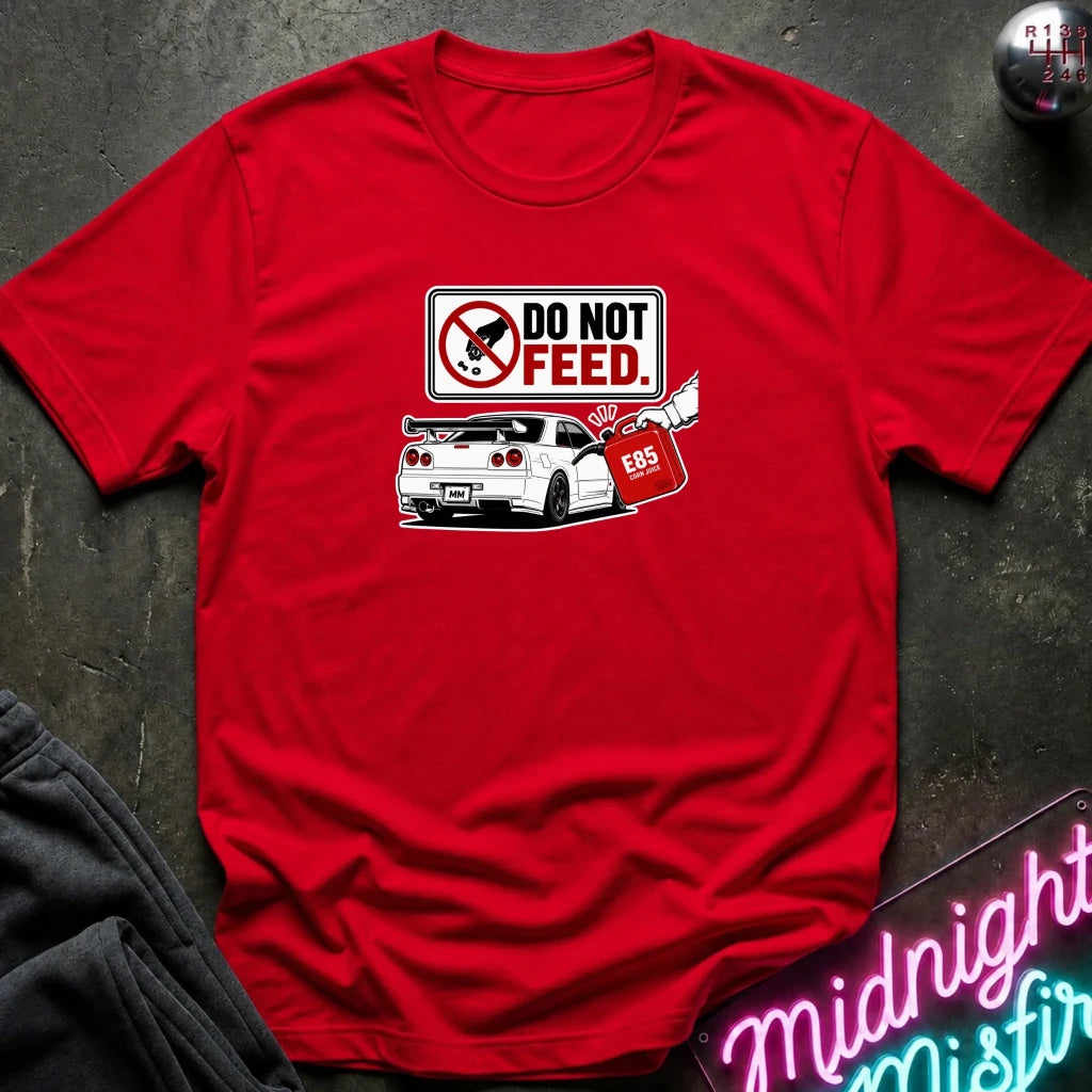 A red short-sleeve crewneck t-shirt laid flat on a dark textured surface. The shirt features a graphic print of a white sports car with a red fuel bag and a 'DO NOT FEED' sign.