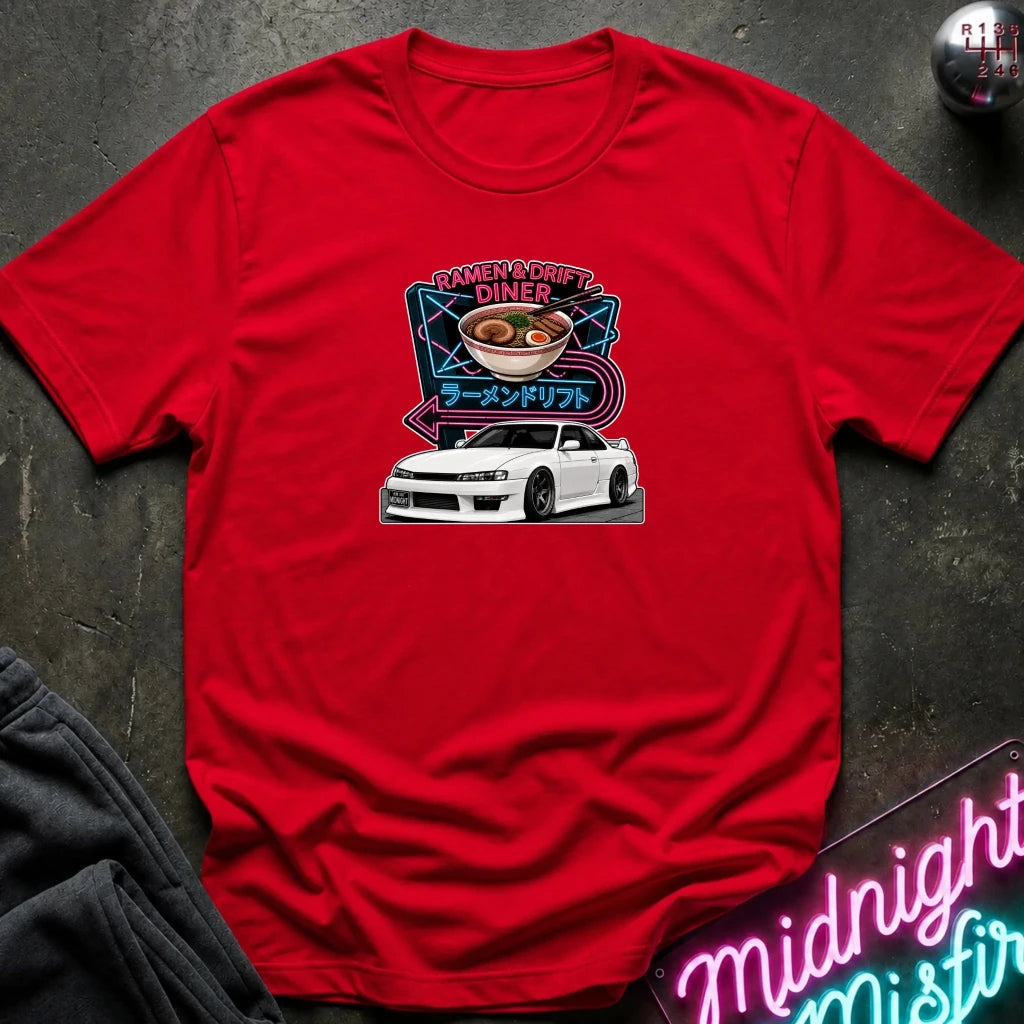 A red short-sleeve graphic t-shirt laid flat on a dark textured surface. The graphic features a white sports car beneath a ramen bowl illustration with neon-style text reading 'Ramen & Drift Diner' and Japanese characters.