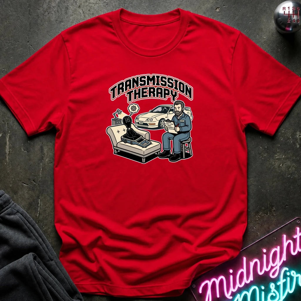 A red short-sleeve graphic T-shirt laid flat on a dark textured surface. The shirt features a cartoon-style print reading 'TRANSMISSION THERAPY' above an illustration of a therapist sitting