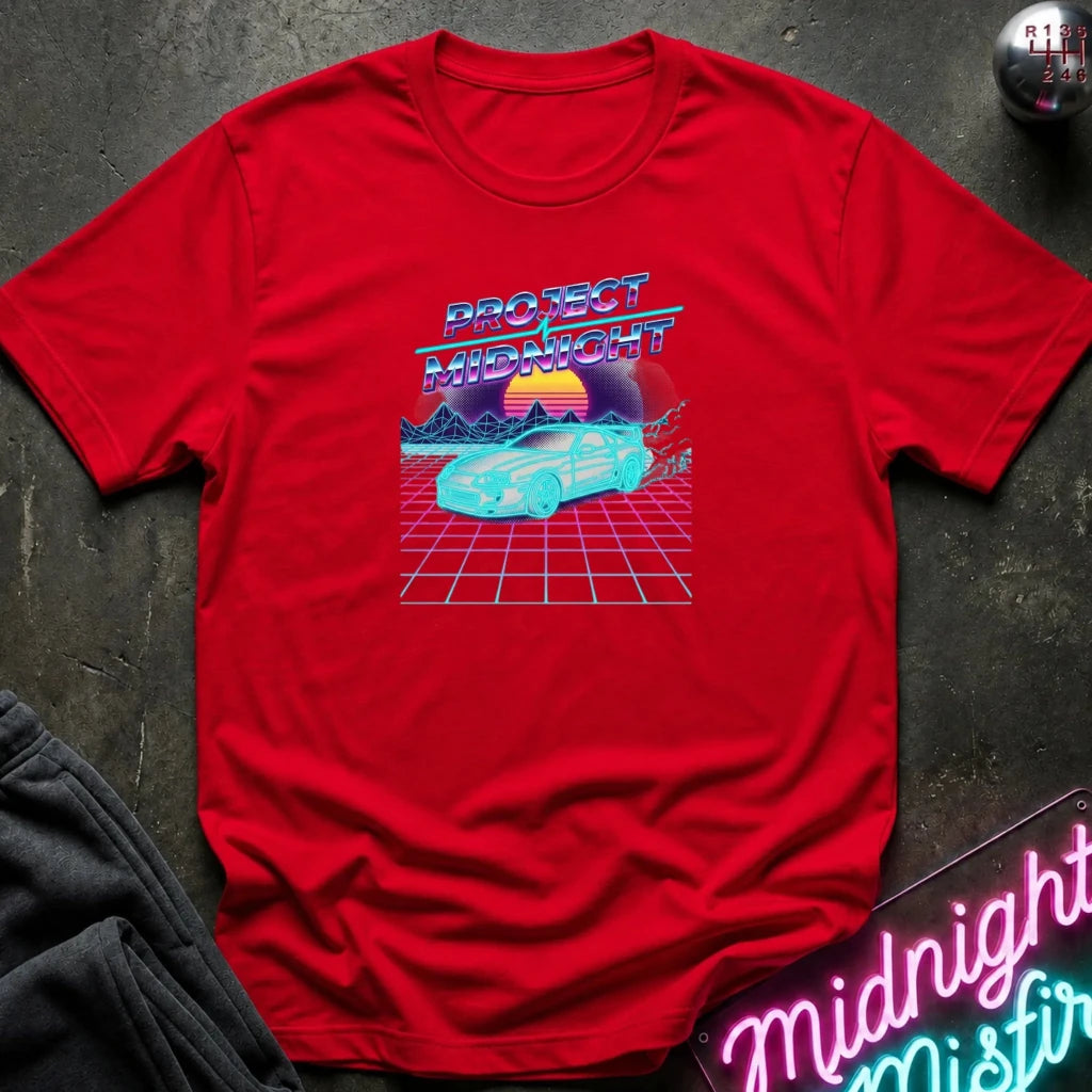 A red short-sleeve t-shirt laid flat on a dark textured surface, featuring a retro 80s-style graphic with the text 'PROJECT MIDNIGHT' above a cyan car on a pink grid with a sunset. A neon sign and folded dark fabric are partially visible around it.