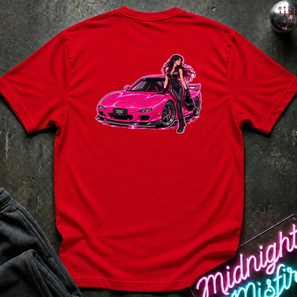 A red short-sleeve t-shirt laid flat on a dark textured surface, featuring a large graphic print of a pink sports car and a woman in black on the back. A gear shift knob is visible in the top right corner, and a neon sign is partially visible in the bottom right.