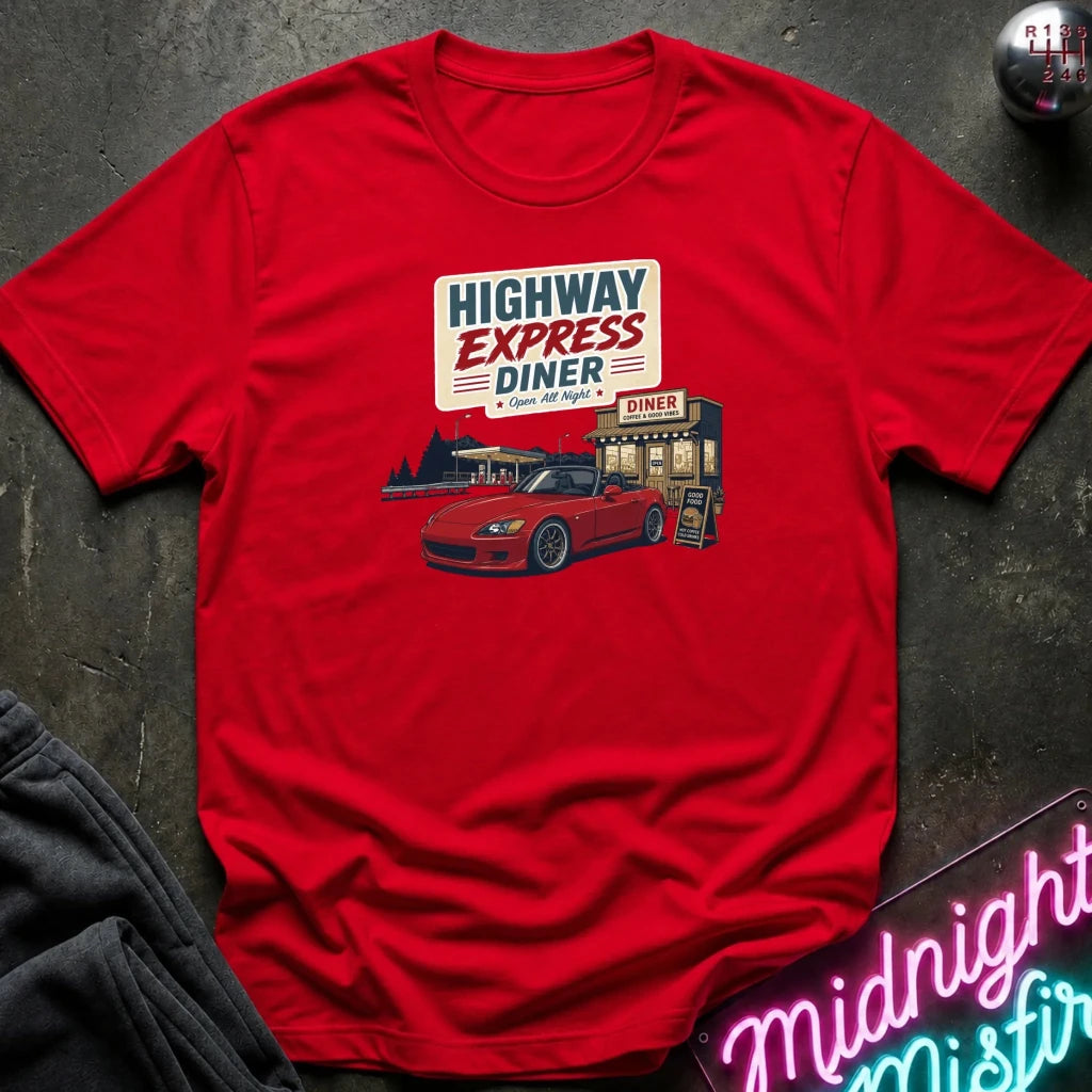 A red short-sleeve graphic t-shirt laid flat on a dark textured surface. The graphic on the chest features a retro-style diner sign reading 'HIGHWAY EXPRESS DINER' above an illustration of a red sports car parked in front of the diner.