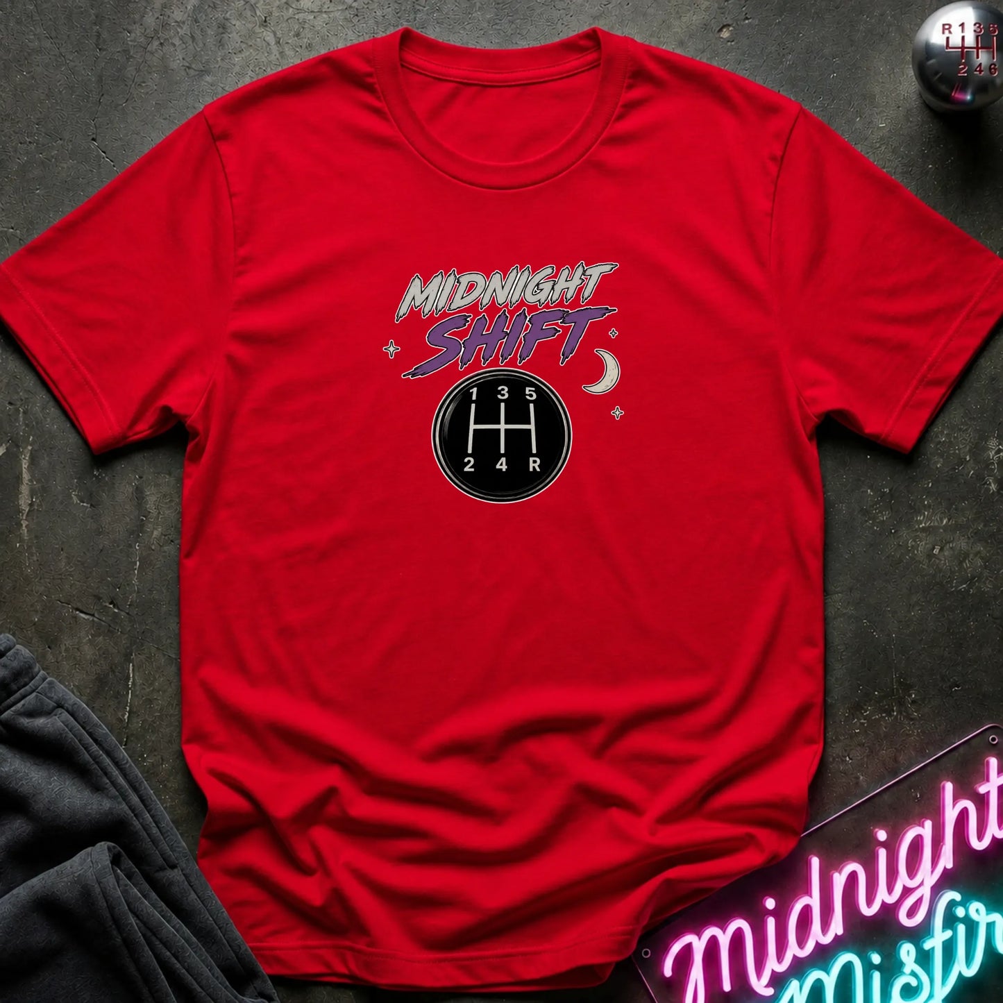A red short-sleeve crewneck t-shirt laid flat on a dark textured surface. The shirt features a graphic on the chest with the words 'MIDNIGHT SHIFT' in stylized white and purple lettering above a black circular gear shift pattern