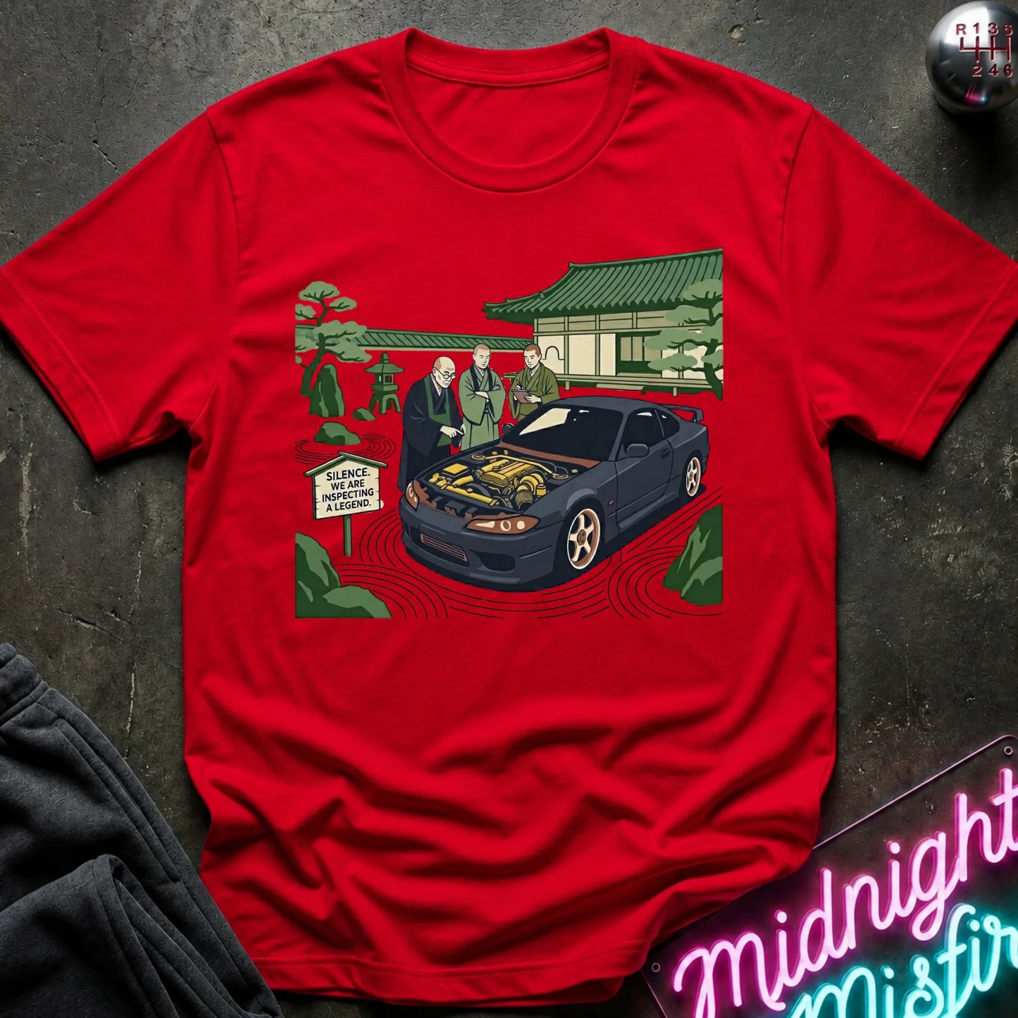 Red short-sleeve graphic t-shirt laid flat on a dark textured surface. The graphic shows a black sports car with its hood open, surrounded by two men in suits, a Japanese-style building, and green trees.