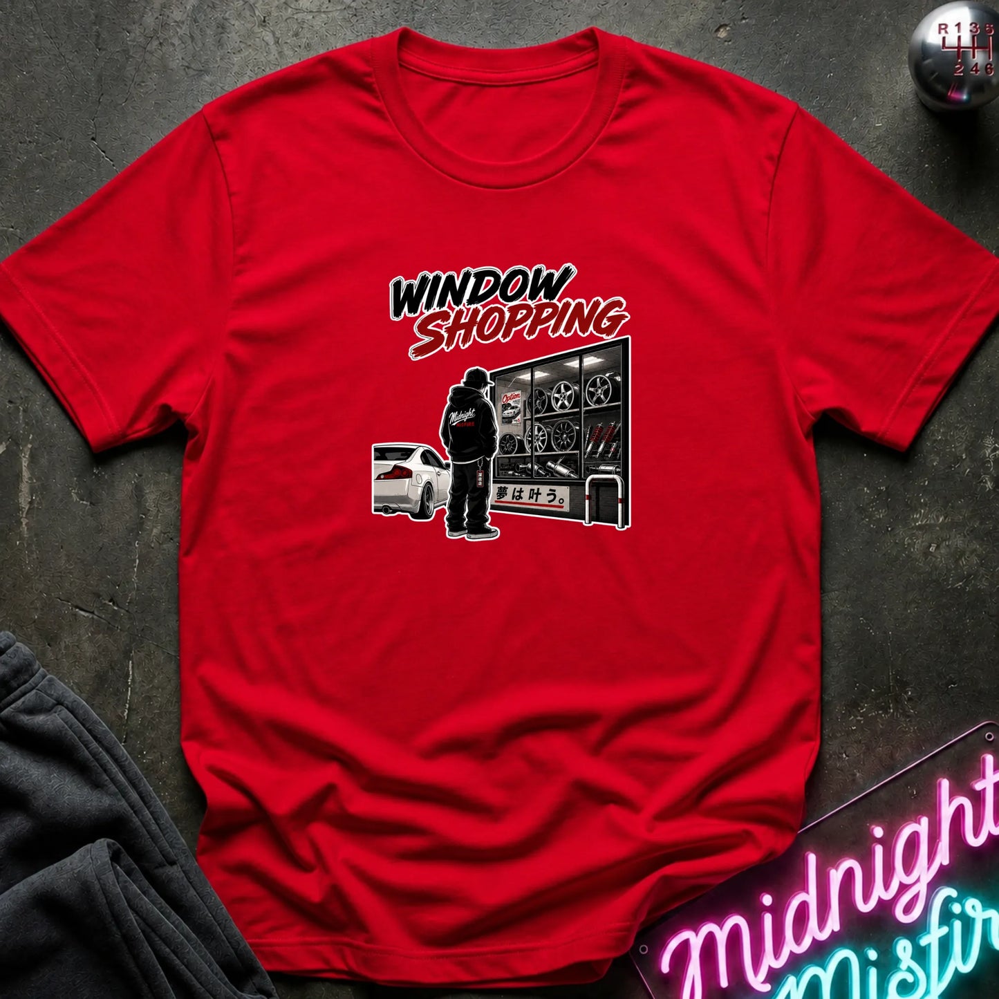 A red short-sleeved t-shirt  that has a cars parked and a person checking out windows of parts.