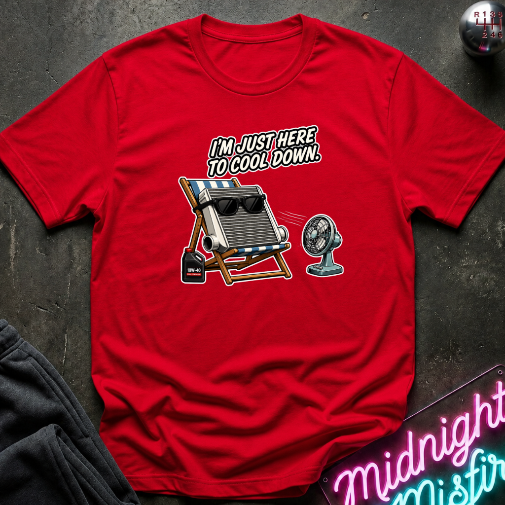 A red short-sleeve graphic t-shirt laid flat on a dark textured surface. The graphic shows a folding beach chair with a radiator on top, a small fan, and the text 'I'M JUST HERE TO COOL DOWN.