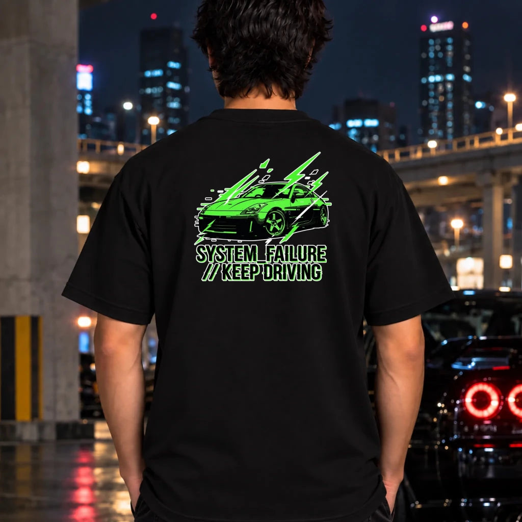 black short-sleeve t-shirt laid flat on a dark textured surface, featuring a graphic of a green sports car with text 'SYSTEM FAILURE // KEEP DRIVING' in green. A neon sign and dark clothing are partially visible as props.