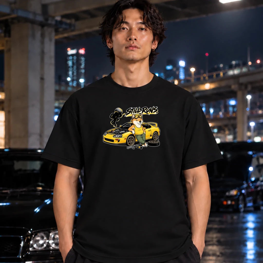 A man wearing a black graphic t-shirt with a cartoon illustration of a yellow sports car and the text 'Still runs' printed on the chest, standing in a nighttime urban setting with blurred cars and city lights in the background.