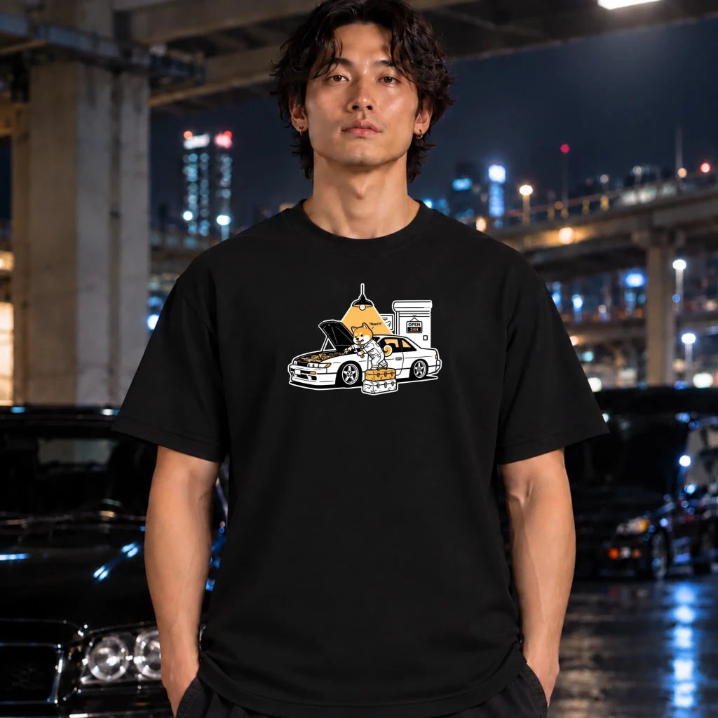 black graphic t-shirt laid flat on a dark textured surface. The graphic features a dog fixing a car.