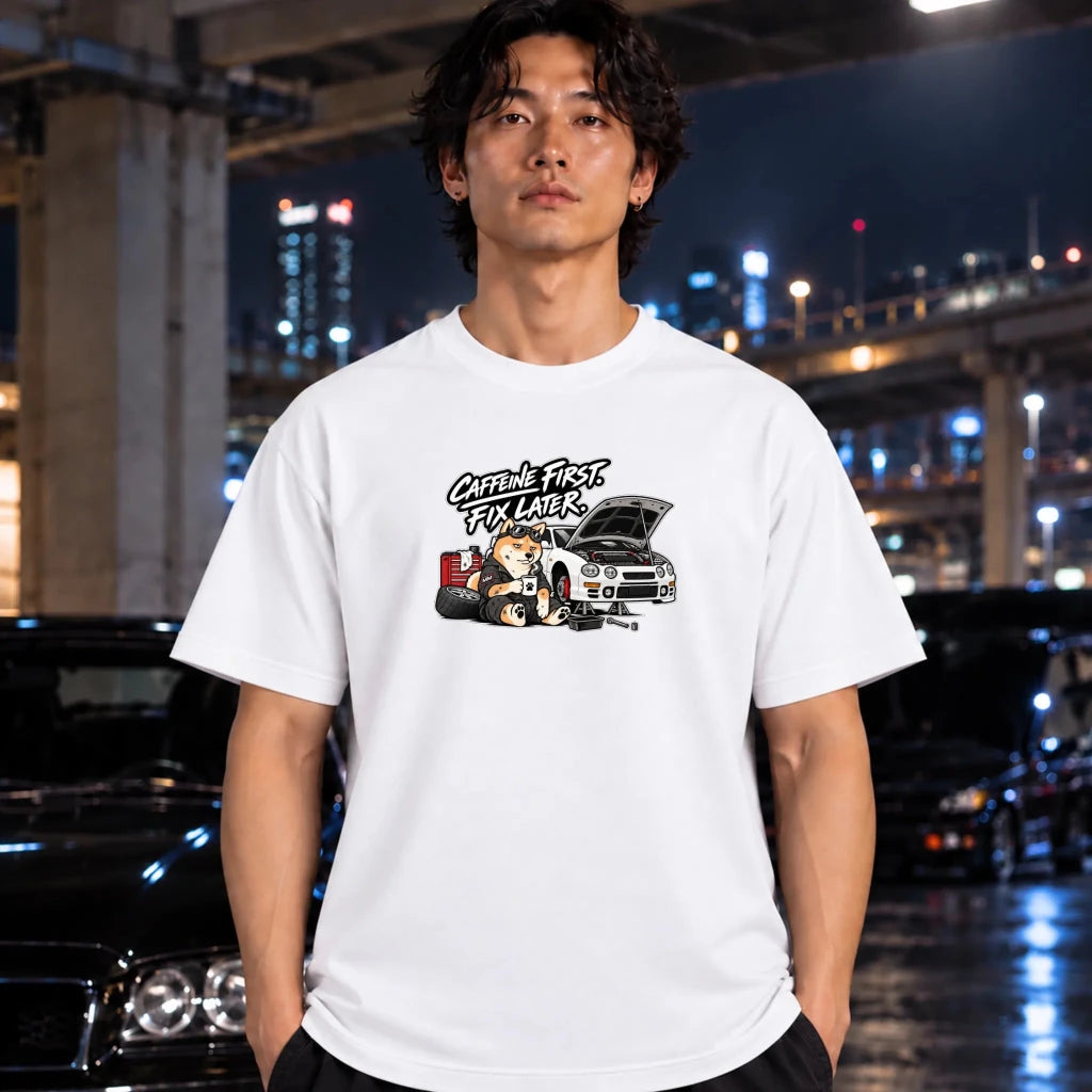 A man wearing a white graphic t-shirt with a 'shiba inu mechanic' car illustration, standing in front of a blurred night city background with cars.