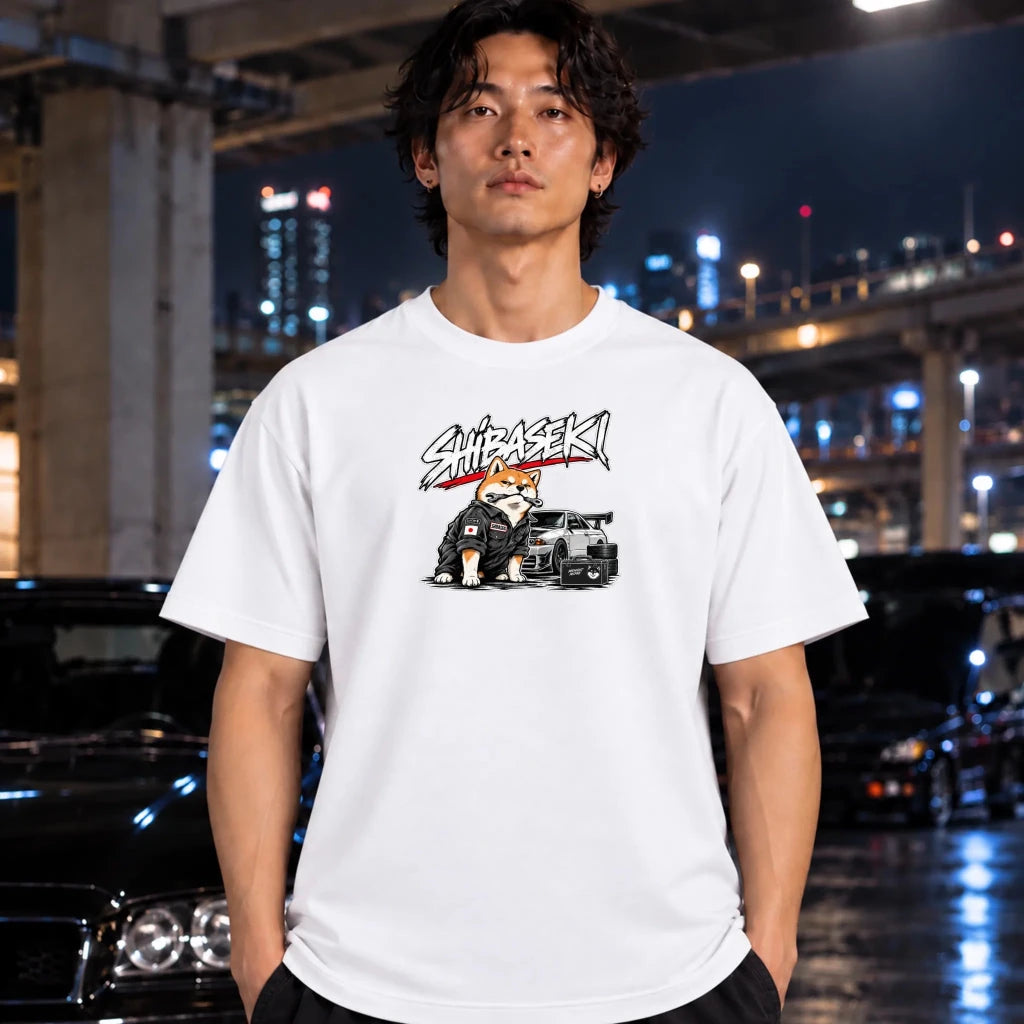 black crewneck t-shirt featuring a graphic of a Shiba Inu dog in a black jacket next to a black sports car, with the text 'SHIBASEKI' printed above. The shirt is worn by a man under the bridge.