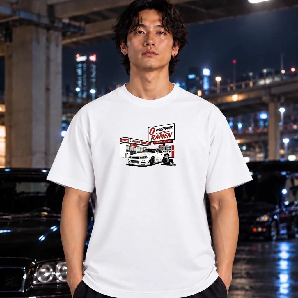 A man wearing a white graphic t-shirt with a retro ramen shop illustration, standing in a city street at night with blurred cars and lights in the background.