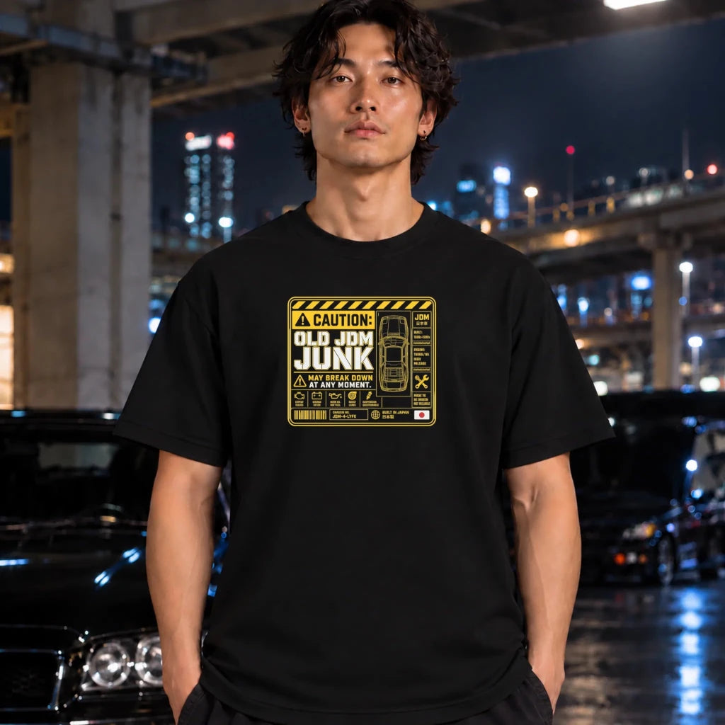 A man wearing a black t-shirt with a yellow and white 'Old JDM Junk' warning label graphic, standing in a nighttime urban setting with blurred cars and city lights in the background.