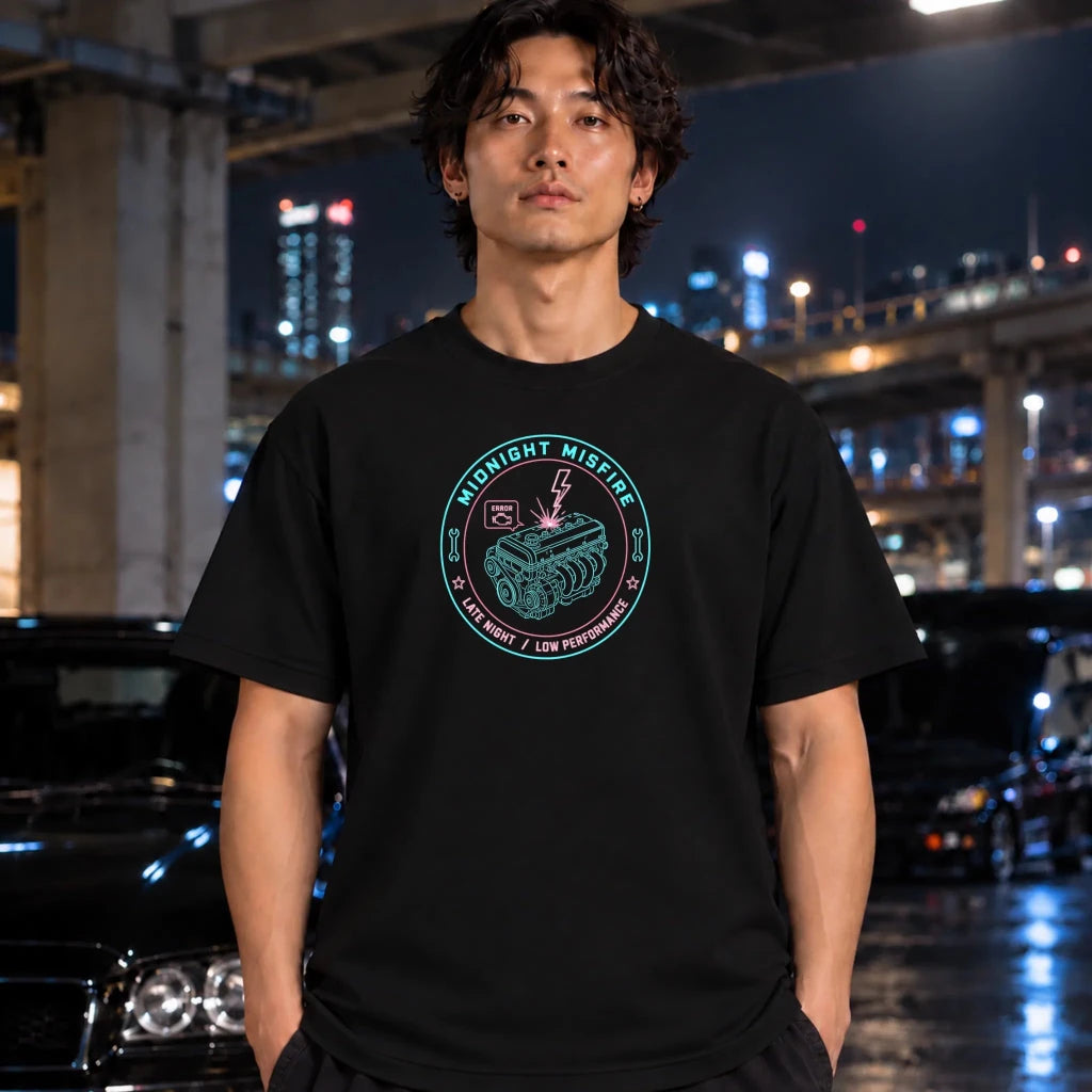 A man wearing a black graphic t-shirt with a circular 'Midnight Misfire' engine design, standing in front of a blurred night city background with cars.