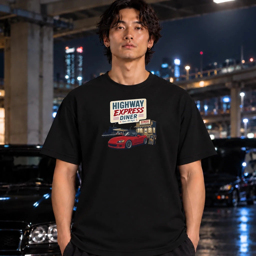A man wearing a black graphic t-shirt with a retro 'Highway Express Diner' sign and red car print, standing in a wet urban night scene with blurred cars and city lights in the background.