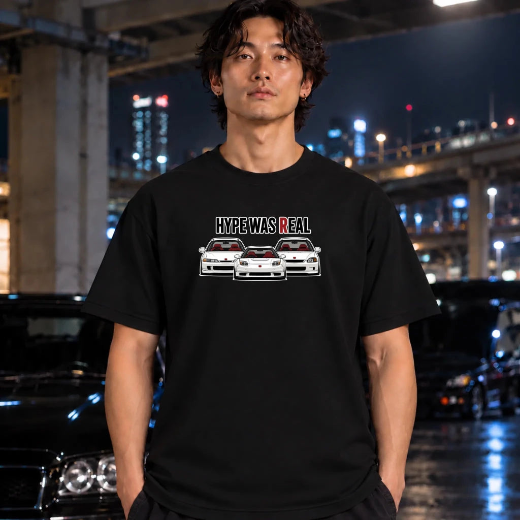 A man wearing a black graphic t-shirt with a 'HYPE WAS REAL' text and three car illustrations, standing in a wet urban night setting with cars and city lights in the background.