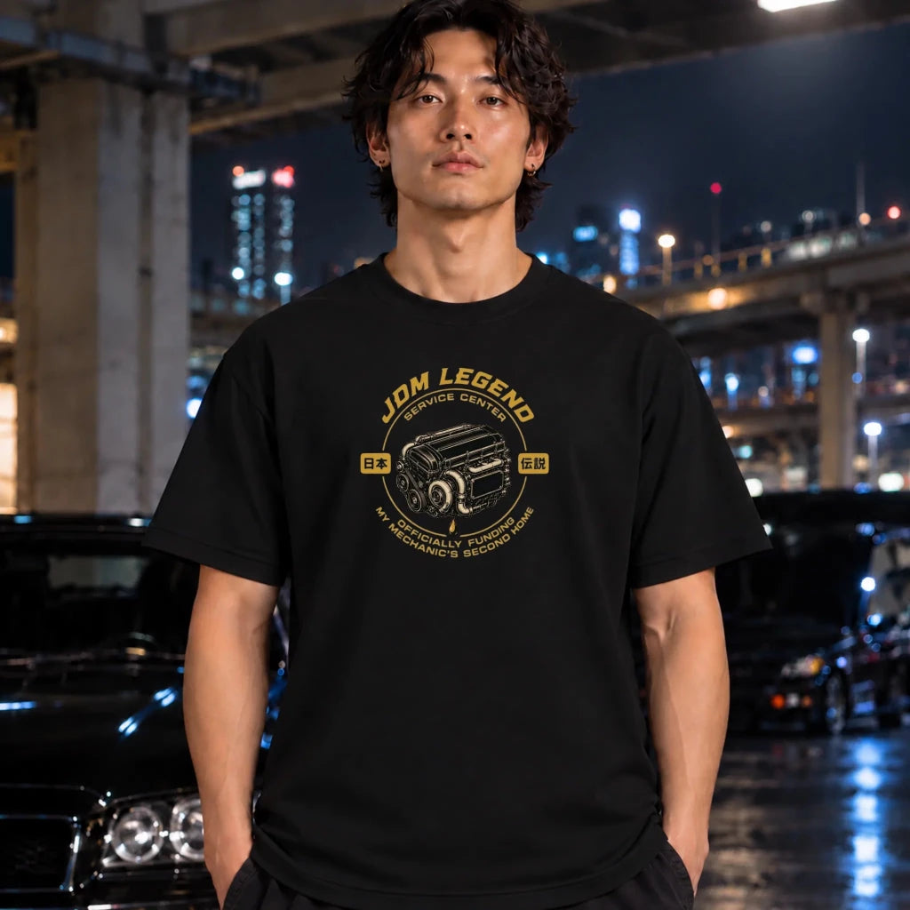 A man wearing a black graphic t-shirt with a yellow 'JDM LEGEND' service center design, standing in front of a blurred night cityscape with cars and lights.