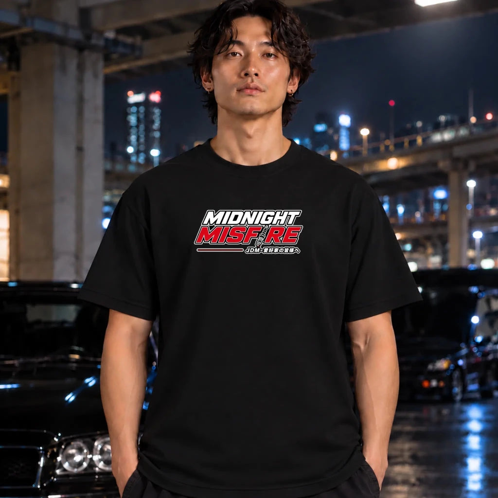 A man wearing a black t-shirt with a 'Midnight Misfire" graphic on the chest, standing in a wet urban parking structure at night with blurred cars and city lights in the background.