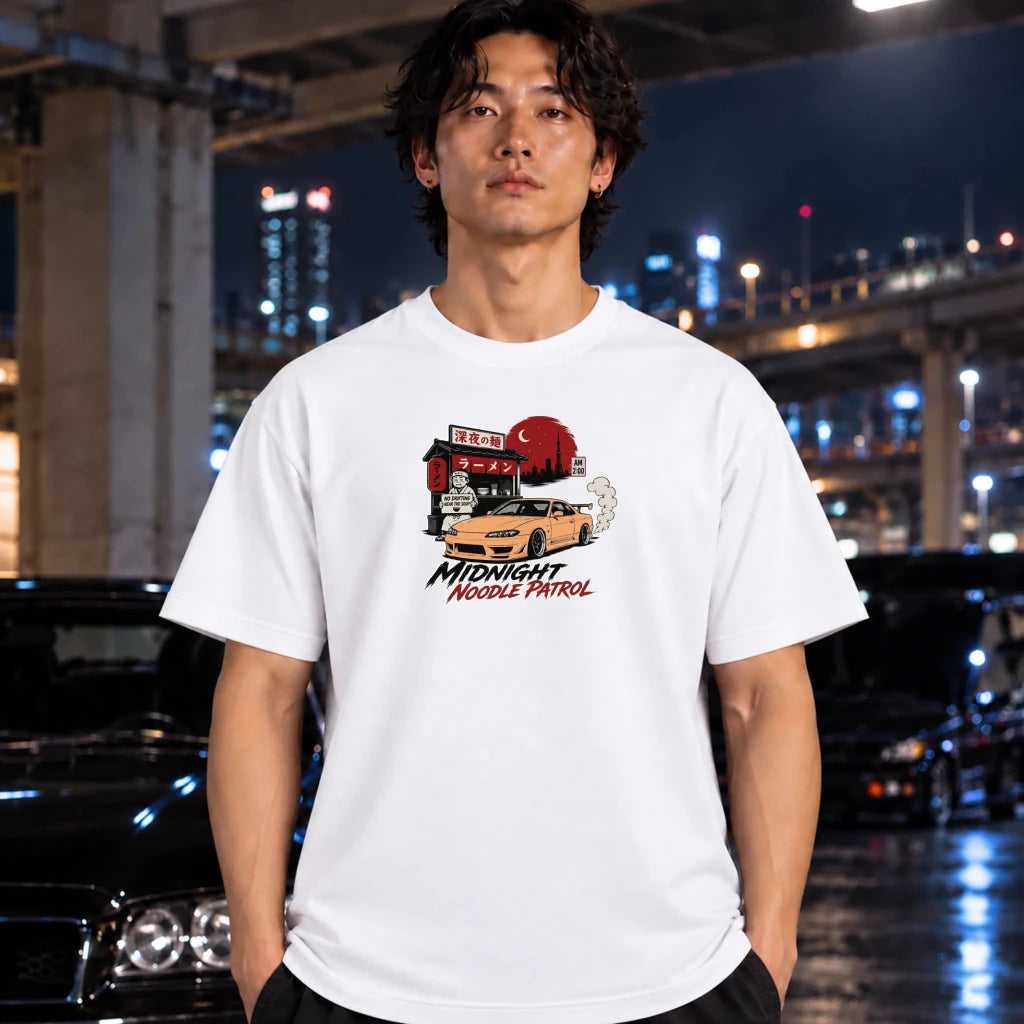 A man wearing a white graphic t-shirt with a 'Midnight Needle Patrol' car illustration, standing in a wet urban street at night with blurred cars and lights in the background.