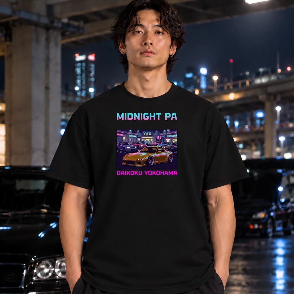 A man wearing a black graphic t-shirt with a 'Midnight Pa' and 'Daikoku Yokohama' print, standing in a wet, neon-lit urban street at night.