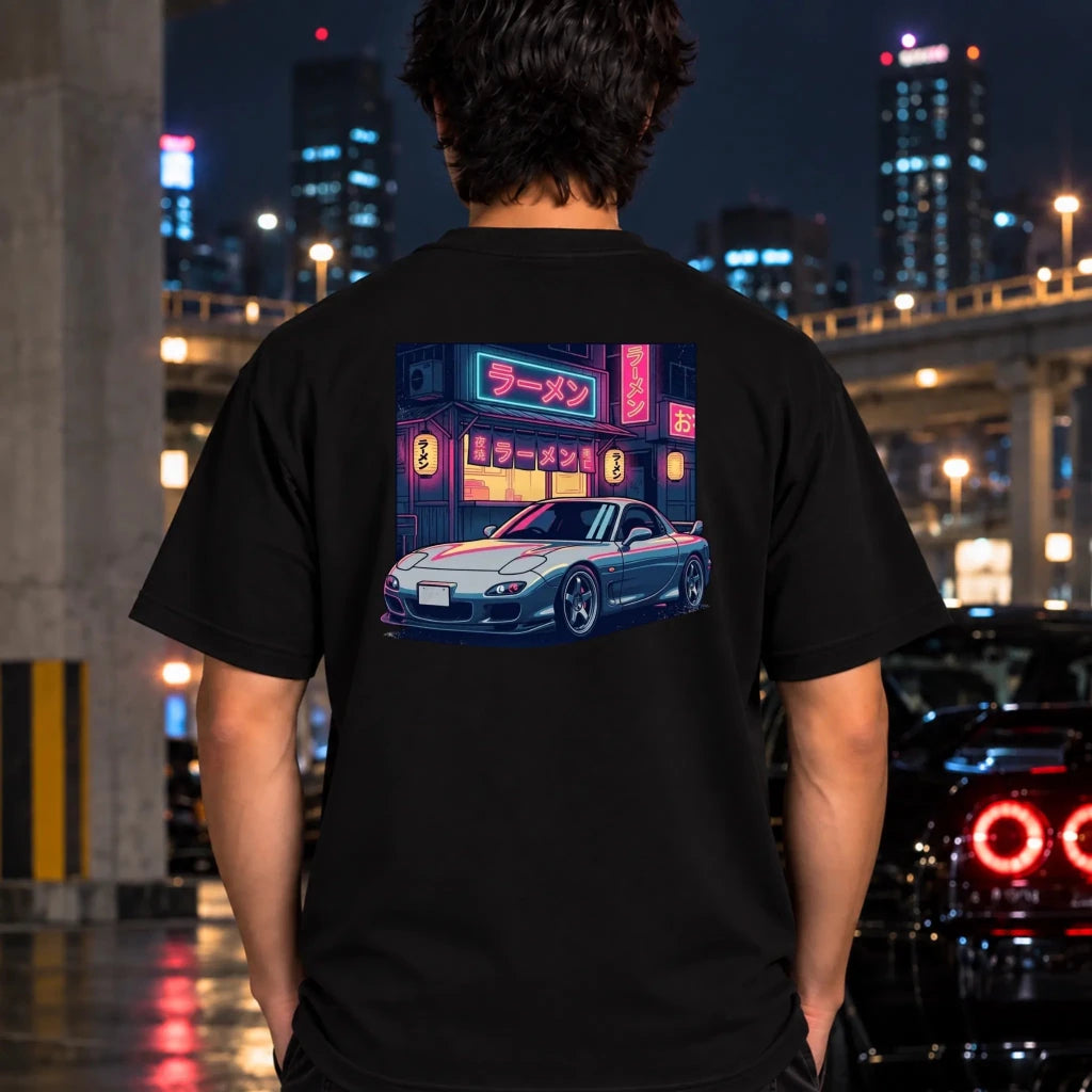 A man viewed from behind wearing a black t-shirt with a large graphic print on the back showing a silver sports car parked in front of a neon-lit ramen shop. The background is a blurred nighttime city street with lights and buildings.