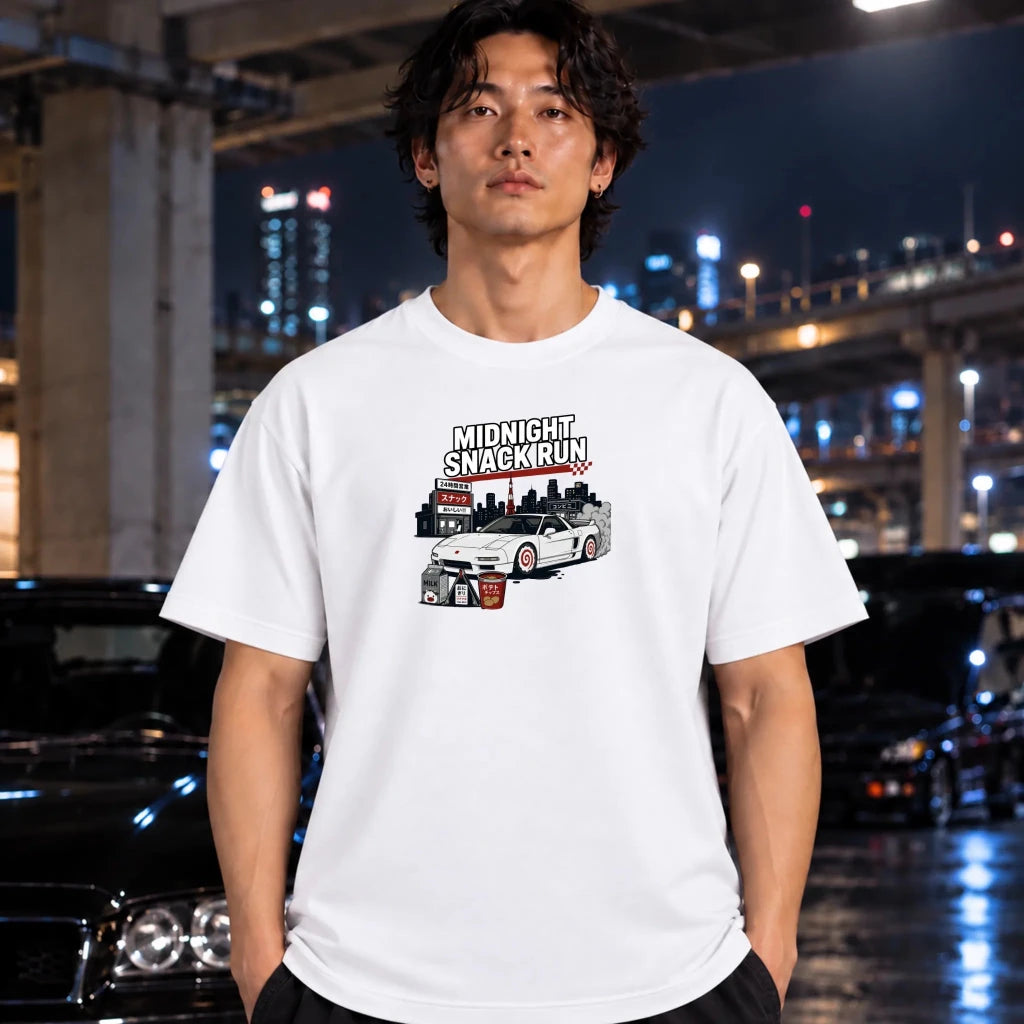 A man wearing a white graphic t-shirt with a 'Midnight Snackrun' car illustration, standing in a city environment at night with blurred cars and lights in the background.