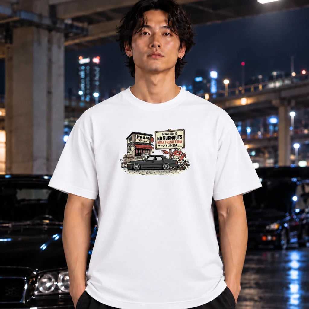 A man wearing a white graphic t-shirt with a vintage-style illustration of a car and a sign that reads 'NO BURNOUTS'. The background is a blurred night city scene with wet pavement reflecting lights.