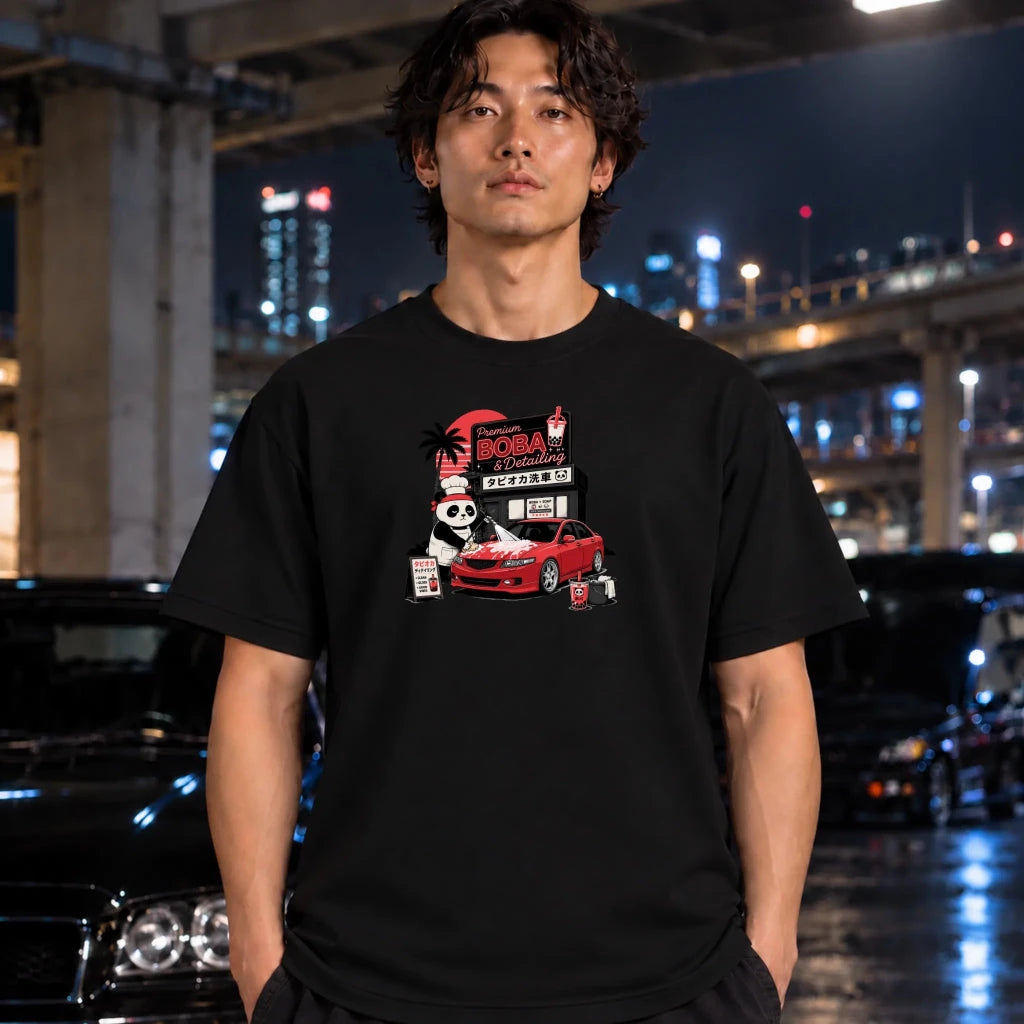 A man wearing a black graphic t-shirt with a 'Boba detailing' cartoon illustration, standing in a wet urban street at night with blurred cars and city lights in the background.