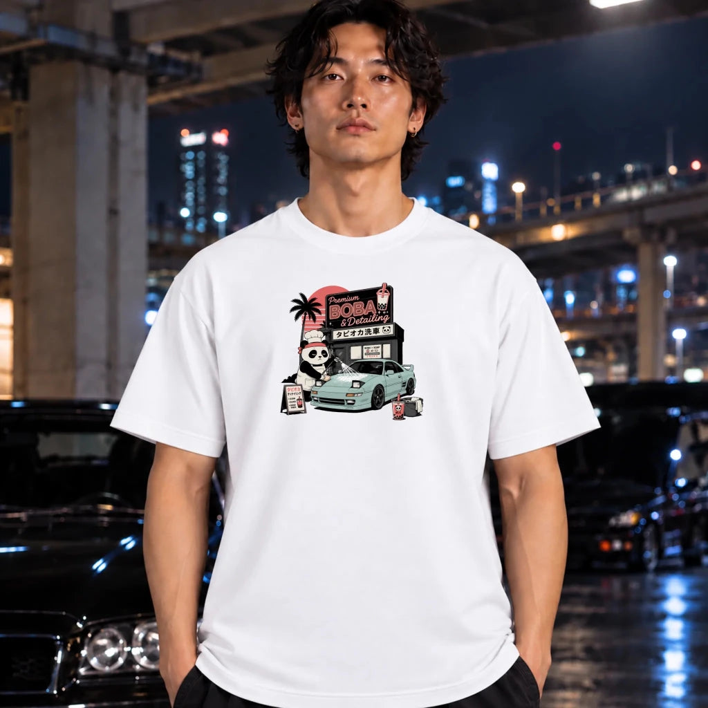 A man wearing a white graphic t-shirt with a cartoon illustration of a light blue sports car and a sign reading 'boba detailing'. The background is a blurred night city scene with wet pavement reflecting lights.