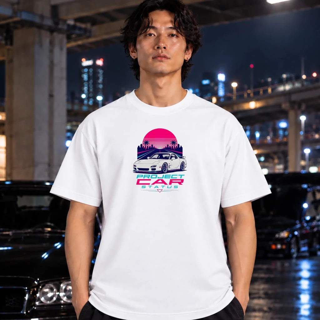 A man wearing a white graphic t-shirt with a retro-futuristic 'Project Car Status' design, standing in a city environment at night with blurred cars and lights in the background.