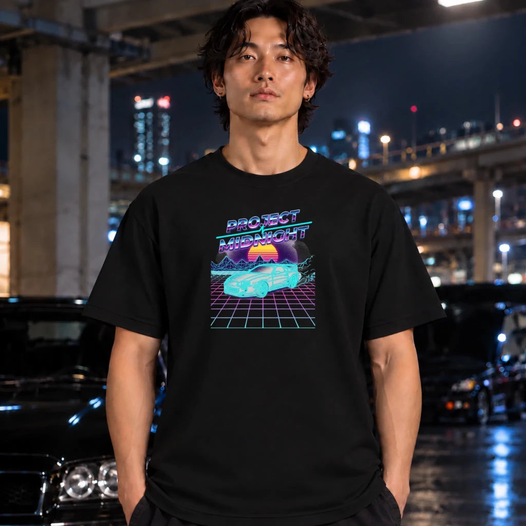 A man wearing a black graphic t-shirt with a retro 80s synthwave design that reads 'PROTECT MiDNIGHT' and features a neon grid and glowing orb graphic, standing in front of a blurred night city background with cars.